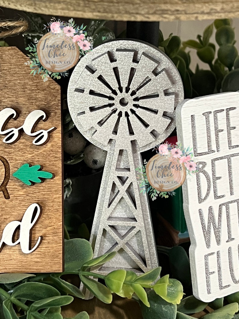 May include: A silver glitter windmill with a wooden base. The windmill is a decorative piece and is surrounded by greenery. The text "Timeless Chic Design Co." is visible on the windmill.