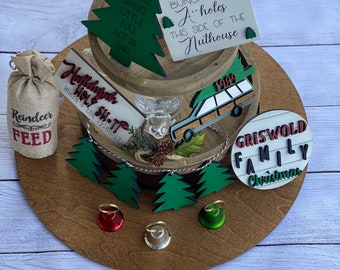 May include: A tiered wooden tray with Christmas-themed decorations. Includes signs with text like "Jolliest bunch of a**holes" and "Griswold Family Christmas." Also features a burlap bag labeled "Reindeer Feed" and miniature Christmas tree cutouts.