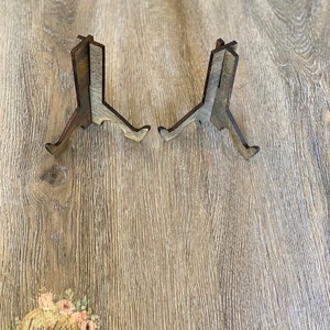 May include: Two wooden display stands with a rustic brown and gray finish. The stands are made of three pieces of wood that are joined together to form a triangular shape. The stands are designed to hold small items, such as photos, cards, or figurines.