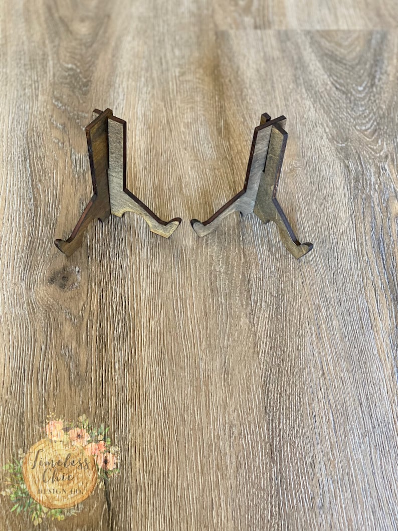 May include: Two wooden display stands with a rustic brown and gray finish. The stands are made of three pieces of wood that are joined together to form a triangular shape. The stands are designed to hold small items, such as photos, cards, or figurines.