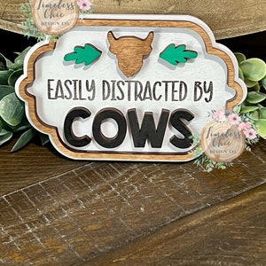 May include: A white wooden sign with a brown cow silhouette and green leaves. The sign reads "Easily Distracted By Cows".