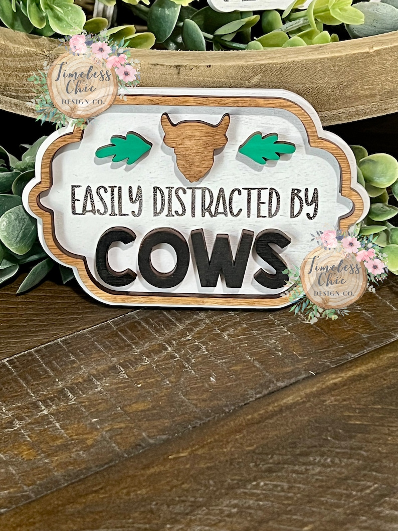 May include: A white wooden sign with a brown cow silhouette and green leaves. The sign reads "Easily Distracted By Cows".