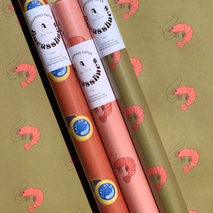 May include: Three rolls of wrapping paper with a shrimp pattern. The rolls are in different colors: green, pink, and orange. The wrapping paper is labeled 'Fine Asslines' and includes the website address 'www.fineasslines.com'. Each roll has a small sticker with the word 'Caviar' and a shrimp design.