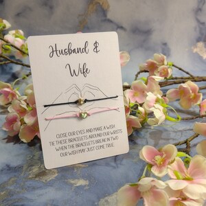 Könnte beinhalten: Eine Karte mit den Worten "Husband & Wife" darauf gedruckt. Zwei Armbänder aus Schnur mit Herzanhängern sind an der Karte befestigt. Die Karte enthält Anweisungen, die Augen zu schließen, einen Wunsch zu machen, die Armbänder um die Handgelenke zu binden, und wenn die Armbänder in zwei Teile zerbrechen, könnte der Wunsch in Erfüllung gehen.