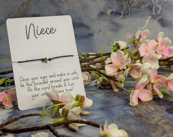May include: A white card with the word "Niece" written on it. A black string bracelet with a gold star charm is attached to the card. The card has the following text: "Close your eyes and make a wish, tie this bracelet around your wrist. As the cord breaks in two, your wish come true!"