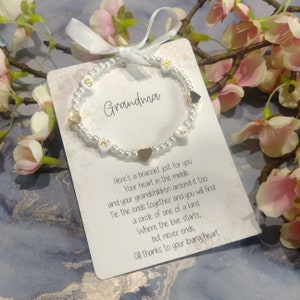 May include: A white pearl bracelet with gold letters spelling "Grandma" and gold heart charms. The bracelet is displayed on a white card with a poem about the love between a grandmother and her grandchildren.