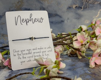 May include: A white card with the word "Nephew" written in black cursive. A black string bracelet with a silver star charm is attached to the card. The card has instructions for making a wish: "Close your eyes and make a wish, tie this bracelet around your wrist. As the cord breaks in two, your wish will come true!"