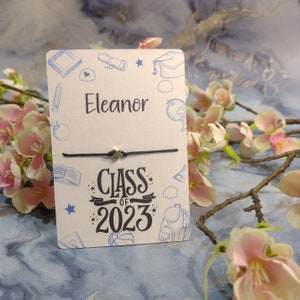 以下が含まれることがあります： 青いインクと黒いインクで描かれた学校用品のイラストと「Class of 2023」の文字が書かれた白いカード。カードには、金色の星形のチャームが付いた黒い紐のブレスレットが付けられています。カードには「Eleanor」という名前も書かれています。