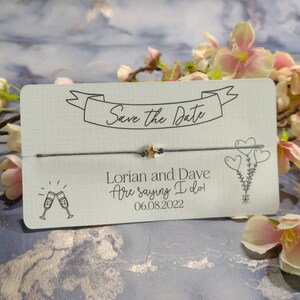 May include: A white card with a gray string tied around it. The card says "Save the Date" and "Lorian and Dave are saying I do" with the date "06.08.2022". There are small gold stars on the string.