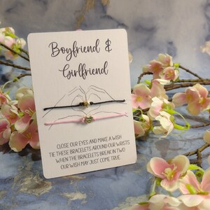 Könnte beinhalten: Eine weiße Karte mit dem Text "Boyfriend & Girlfriend" darauf gedruckt. Zwei Armbänder aus Schnur mit goldenen Anhängern sind an der Karte befestigt. Die Karte liegt auf einem Hintergrund aus rosa Blumen.