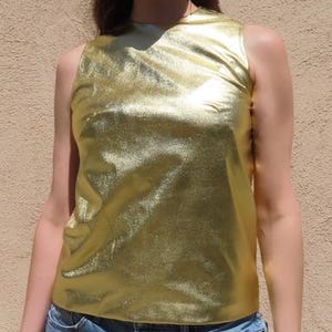 May include: A sleeveless gold metallic top paired with denim shorts. The top has a textured, shiny appearance. The person is wearing green-tinted sunglasses.