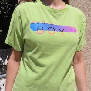 May include: A light green t-shirt with the text "Boogie Boy" in a colorful, brushstroke design. The shirt is a crew neck style and is paired with blue jeans. The person is wearing heart-shaped sunglasses.