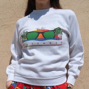 Pode incluir: Uma pessoa vestindo um moletom branco com um gr&aacute;fico de &oacute;culos de sol e o texto "Ray-Ban Apparel". A pessoa tamb&eacute;m est&aacute; usando &oacute;culos de sol vermelhos e cal&ccedil;as estampadas vermelhas.