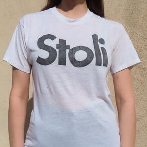 以下が含まれることがあります： 「Stoli」の文字が濃いグレーでプリントされた白いTシャツ。Tシャツは半袖で、クルーネックです。画像に写っている人物は黒いパンツと赤いサングラスを着用しています。