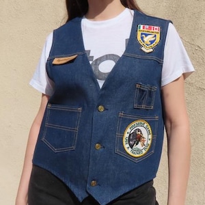 May include: A dark blue denim vest with multiple patches and gold buttons. The vest has two front pockets and a small leather tag. The patches feature various designs, including a bird and a horse. The person is wearing a white t-shirt underneath.