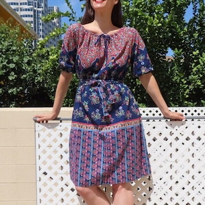 May include: A vintage knee-length dress with a floral pattern in blue, red, and white. The dress has short sleeves, a tie-neck, and a matching belt. The model is wearing brown sandals and red sunglasses. The dress is approximately 100 cm in length.