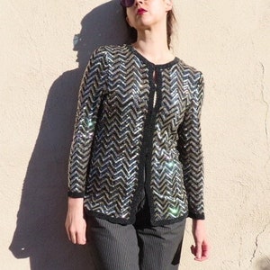 May include: A sequined jacket with a black and silver chevron pattern. The jacket has a black trim and is paired with gray pinstripe pants. The person in the image is wearing sunglasses and leaning against a beige wall.