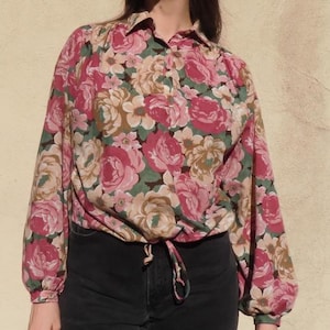 Puede incluir: Una blusa con estampado floral con mangas largas y un lazo en la cintura. La blusa presenta un patrón de rosas rosas y crema con hojas verdes sobre un fondo apagado. La modelo lleva pantalones negros y gafas de sol.