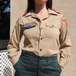 Puede incluir: Una camisa beige con parches y el texto "Boy Scouts of America". La camisa tiene dos bolsillos en el pecho y se combina con pantalones verde oscuro. El atuendo recuerda a un uniforme de explorador vintage.
