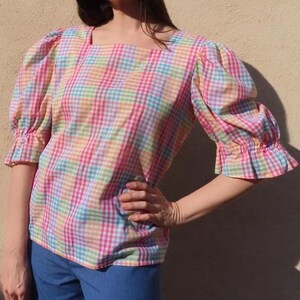 May include: A colorful plaid blouse with a square neckline and three-quarter sleeves. The blouse features a pastel rainbow pattern of pink, yellow, blue, and green checks. The sleeves have ruffled cuffs. The garment is made of a lightweight fabric.