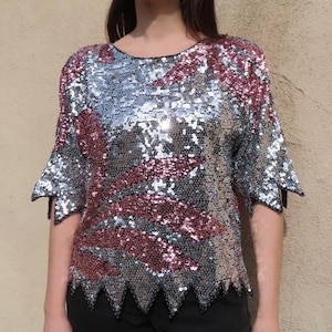 Può includere: Top di paillettes argento e bordeaux con maniche corte e orlo smerlato. Il top ha uno scollo tondo ed è impreziosito da paillettes scintillanti. Le maniche hanno un design svasato. Il top è abbinato a pantaloni neri.