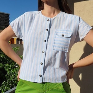 Puede incluir: Camisa de manga corta a rayas azul claro y blanco con un bolsillo en el pecho. La camisa tiene una botonadura frontal y se combina con pantalones verdes brillantes. El atuendo se usa al aire libre en un día soleado.