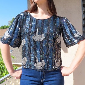 May include: A sequined top with a round neckline and short sleeves. The top features a pattern of vertical stripes in black and blue, with silver embellishments. The garment is paired with blue jeans.