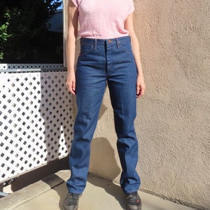 May include: A person wearing a light pink V-neck shirt and dark blue denim jeans. The jeans have a high waist and a straight leg. The person is wearing dark sneakers and pink sunglasses.