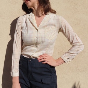 May include: A cream-colored, long-sleeved shirt with a plaid pattern in red and blue. The shirt has a collared neckline, a single chest pocket, and button closures. The model is wearing the shirt with dark blue pants.