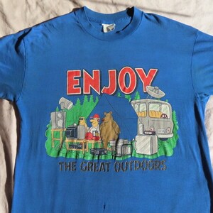 May include: Blue t-shirt with a camping-themed graphic. The graphic features the word "ENJOY" in red, cartoon bears, a tent, a camper van, and the text "THE GREAT OUTDOORS".