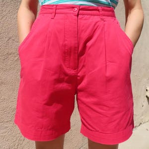 May include: Bright red shorts with pleats and pockets. The shorts are knee-length and have a button closure. The person in the image is wearing a striped top. The shorts are made of a solid, opaque fabric.