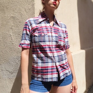 May include: A short-sleeved plaid shirt in red, white, and blue. The shirt has two chest pockets and a button-down front. The model is wearing blue denim shorts and red sunglasses. The background is a beige stucco wall.
