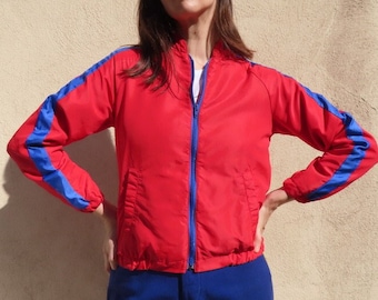 May include: A red jacket with blue stripes on the sleeves. The jacket has a zipper closure and is worn with blue pants. The jacket is a vibrant red color and the blue stripes add a contrasting detail.