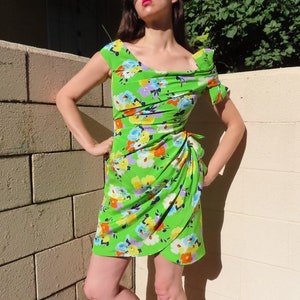 Puede incluir: Un vestido verde brillante con estampado floral, con un escote drapeado y una falda cruzada. El vestido presenta un patrón vibrante de flores amarillas, azules y moradas. El vestido es hasta la rodilla y tiene un detalle de lazo en la cintura.