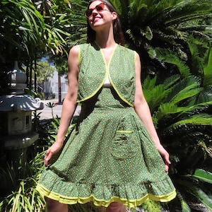 May include: A vintage-style green dress and matching vest set with a small white and brown floral pattern. The dress features a ruffled hem and a pocket, both trimmed with bright yellow. The vest has a yellow trim as well.