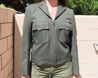 May include: An olive green jacket with a zip-up front, two chest pockets with gold-colored button closures, and shoulder epaulets. The jacket is paired with olive green pants. The background features a brick wall and green foliage.