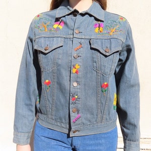 May include: A light blue denim jacket with colorful embroidered floral and insect designs. The jacket features a classic button-up front, two chest pockets, and a collared neckline. The embroidery includes red, yellow, and green accents.