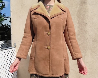 May include: A tan suede coat with a shearling collar and lining. The coat has two large buttons and two front pockets. The coat is knee-length and has a classic, vintage style.