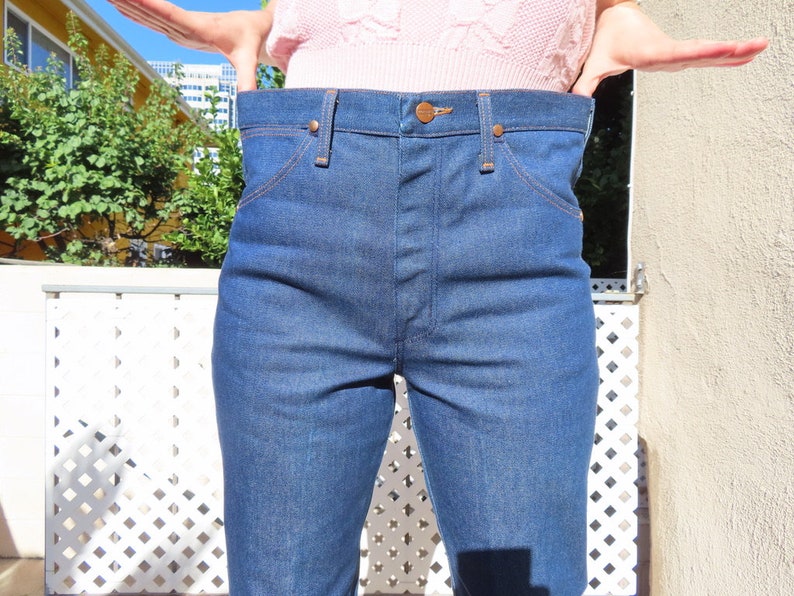 May include: Blue denim jeans with a high waist and classic five-pocket design. The jeans have copper-colored rivets and stitching. The denim is a medium wash, and the jeans appear to be unworn.