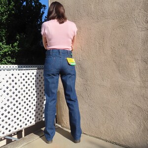 May include: Dark blue denim jeans with a high waist and a straight leg. The jeans have a classic five-pocket design and a yellow tag on the back pocket. The person is wearing a light pink short-sleeved top.