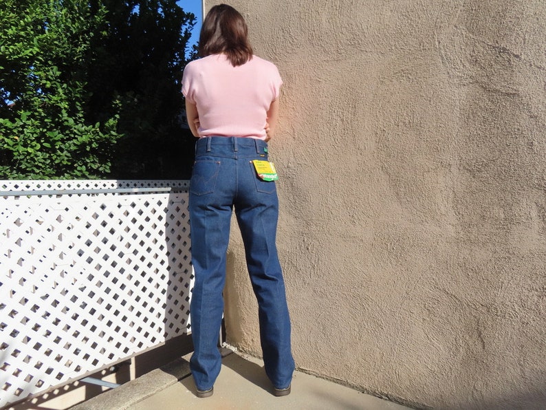 May include: Dark blue denim jeans with a high waist and a straight leg. The jeans have a classic five-pocket design and a yellow tag on the back pocket. The person is wearing a light pink short-sleeved top.