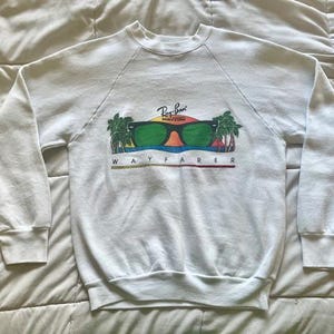 Pode incluir: Camisola branca com gola redonda e mangas compridas. A frente apresenta um gr&aacute;fico de &oacute;culos de sol verdes com o log&oacute;tipo Ray-Ban, palmeiras e a palavra "WAYFARER" em letras coloridas.