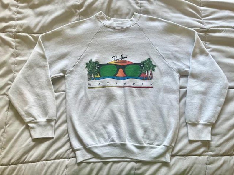 Pode incluir: Camisola branca com gola redonda e mangas compridas. A frente apresenta um gr&aacute;fico de &oacute;culos de sol verdes com o log&oacute;tipo Ray-Ban, palmeiras e a palavra "WAYFARER" em letras coloridas.