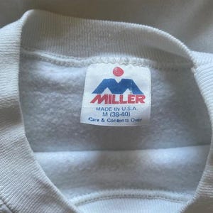 Pode incluir: Close-up de um moletom branco com gola redonda. A etiqueta diz "MILLER" em vermelho e azul, com "MADE IN U.S.A." e o tamanho M (38-40) impressos abaixo. As instru&ccedil;&otilde;es de cuidado tamb&eacute;m est&atilde;o impressas na etiqueta.