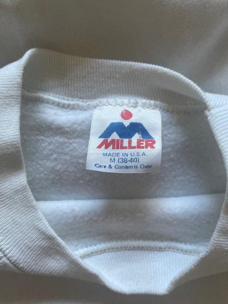 Pode incluir: Close-up de um moletom branco com gola redonda. A etiqueta diz "MILLER" em vermelho e azul, com "MADE IN U.S.A." e o tamanho M (38-40) impressos abaixo. As instru&ccedil;&otilde;es de cuidado tamb&eacute;m est&atilde;o impressas na etiqueta.