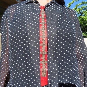 May include: A sheer black blouse with white polka dots. A vertical red ribbon with ornate gold embellishments runs down the center. The blouse has a collared neckline and buttoned front.