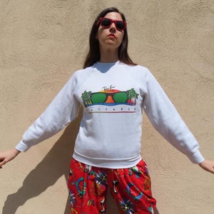 Pode incluir: Uma camisola branca com um gr&aacute;fico Ray-Ban Wayfarer com &oacute;culos de sol, palmeiras e a palavra "WAYFARER" em cores do arco-&iacute;ris. A pessoa est&aacute; usando &oacute;culos de sol vermelhos e cal&ccedil;as estampadas vermelhas.