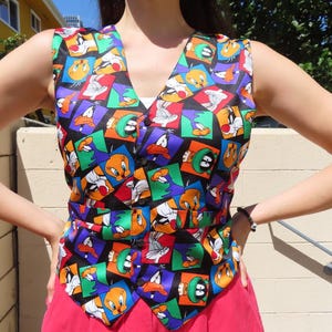 May include: A colorful vest featuring cartoon characters in a repeating pattern. The vest has a V-neck and a button closure. The background is black, and the characters are in squares of various colors.