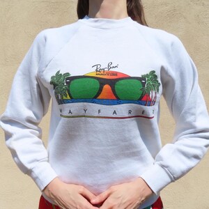 Pode incluir: Camisola branca com um gr&aacute;fico colorido de &oacute;culos de sol, um p&ocirc;r do sol e palmeiras. O texto "Ray-Ban Bausch & Lomb WAYFARER?" est&aacute; impresso abaixo. A camisola tem mangas compridas e gola redonda.