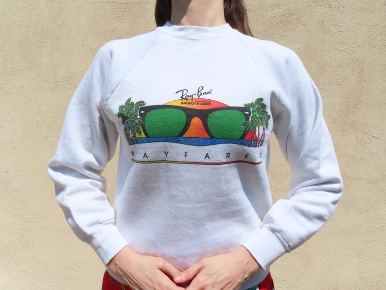 Pode incluir: Camisola branca com um gr&aacute;fico colorido de &oacute;culos de sol, um p&ocirc;r do sol e palmeiras. O texto "Ray-Ban Bausch & Lomb WAYFARER?" est&aacute; impresso abaixo. A camisola tem mangas compridas e gola redonda.